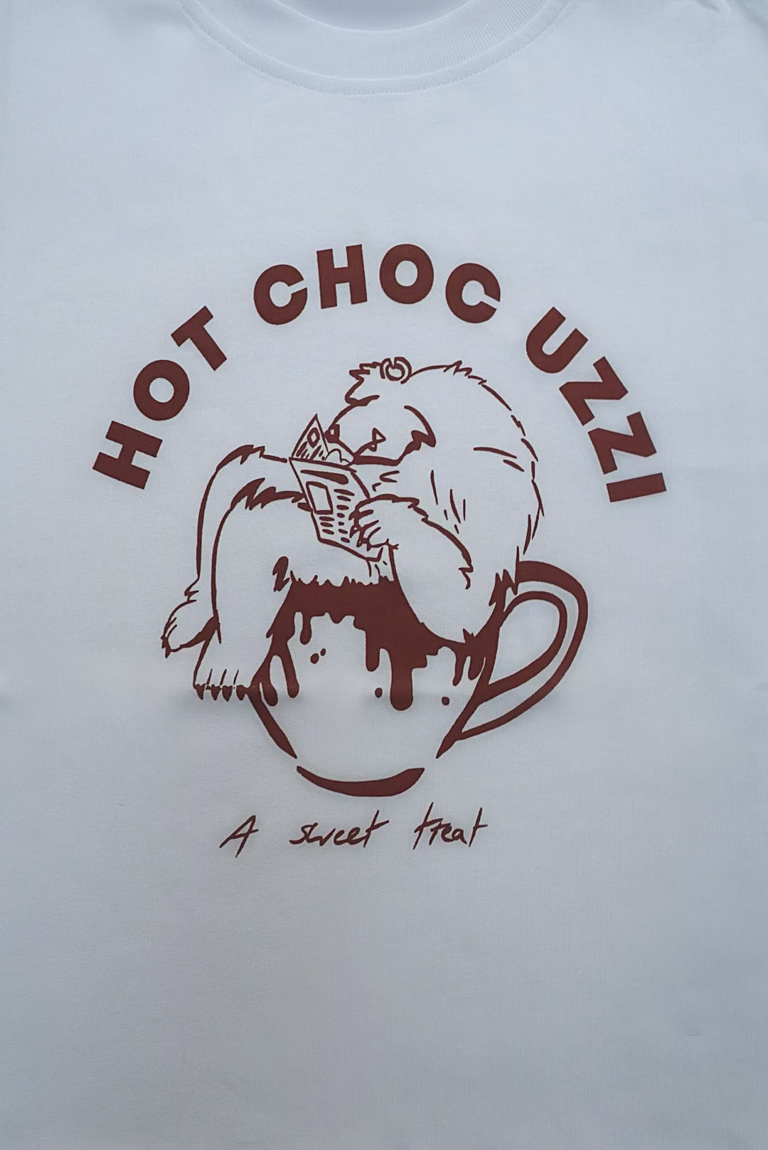 White T-shirt with cartoon animal lounging in a mug, labeled HOT CHOC UZZI and A sweet treat, perfect for winter coziness.