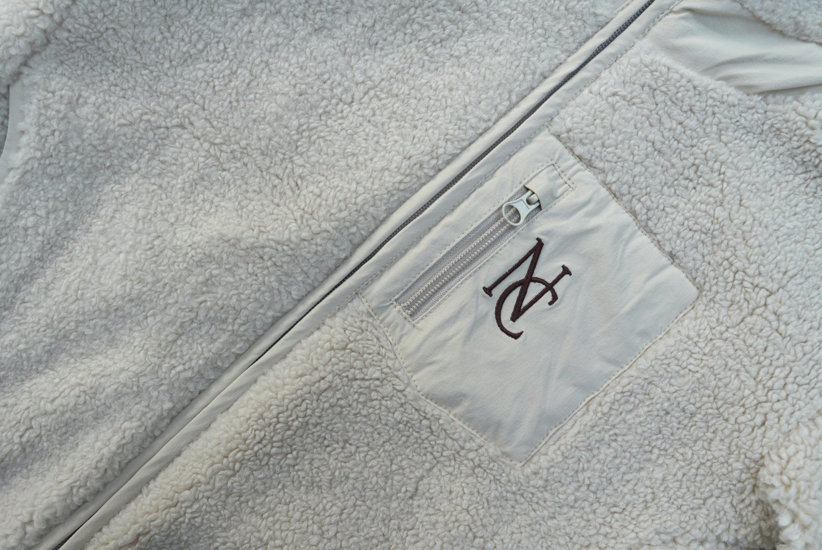 Close-up of the Cosy Teddy Jacket, featuring plush sherpa-fleece, a vertical chest zip-pocket, and an embroidered “NC” monogram on the pocket panel.