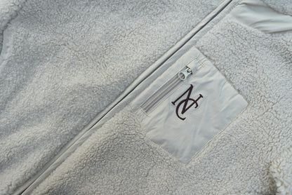 Close-up of the Cosy Teddy Jacket, featuring plush sherpa-fleece, a vertical chest zip-pocket, and an embroidered “NC” monogram on the pocket panel.